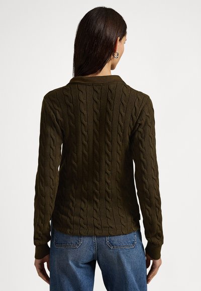 Γυναίκα με μακριά σκούρα μαλλιά, φοράει σκούρο πράσινο πουλόβερ με πλέξη καλώδιο και μπλε τζιν, φαίνεται από την πίσω πλευρά με φόντο λευκό.