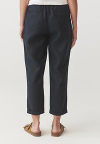Persoon met zwarte cropped broek met omgeslagen pijpen en bruine instappers, gezien van achteren tegen een effen achtergrond.