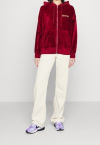 Felpa rossa in velluto con zip, coulisse e tasche frontali; abbinata a pantaloni larghi crema e scarpe sportive viola con accenti grafici.