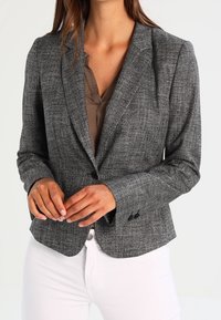 Blazer à carreaux gris au design ajusté, avec des revers notés, fermeture à un bouton, et tissu texturé. Assorti à un chemisier clair et un pantalon blanc.