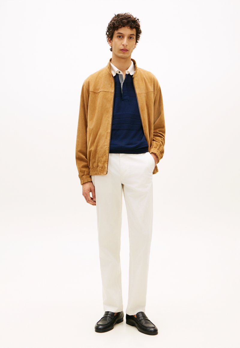 Ταν σαμουαρέ μπουφάν bomber, μπλε σκούρο πόλο πουκάμισο με τρία κουμπιά, λευκό παντελόνι ίσιας γραμμής και μαύρα παπούτσια χωρίς κορδόνια. Μινιμαλιστικός σχεδιασμός.