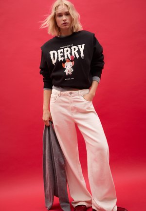Svart sweatshirt med texten "WELCOME TO DERRY" och en figur som håller en röd ballong. Matchad med ljusrosa vida byxor.