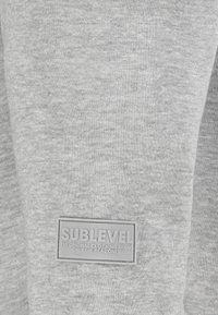 Graues Sweatshirt-Material mit einer weichen Textur. Enthält ein rechteckiges Gummilogo-Patch mit der Prägung "SUBLEVEL" und zusätzlichem Text.