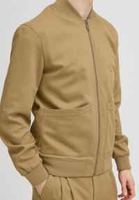 Tan zip-up bomberjack gemaakt van textuurstof, met zijvakken, geribbelde manchetten en een bomberkraag ontwerp.