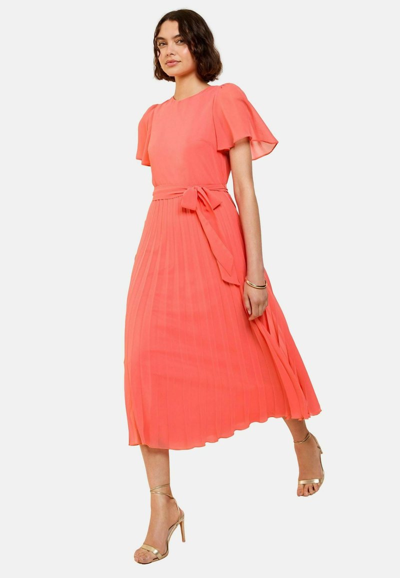 Koralfarbenes plissiertes Midikleid mit kurzen schwingenden Ärmeln und einem Taillengurt. Glatter Stoff und ein ausgestellter Rock. Getragen mit goldenen Riemchenheels.