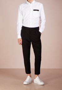 Weißes Langarm-Hemd mit Knopfleiste und schwarzem Taschen-Detail, kombiniert mit schwarzen, maßgeschneiderten Hosen und weißen Sneakers, vor einem hellen Hintergrund stehend.