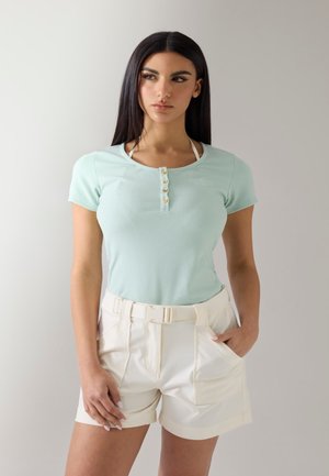 Jeune femme aux longs cheveux foncés portant un haut côtelé vert menthe à manches courtes et un short beige taille haute avec ceinture.