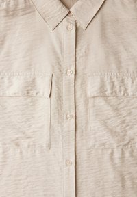 Camicia beige con bottoni realizzata in tessuto leggero, con una finitura testurizzata, due tasche anteriori e un classico design del colletto.
