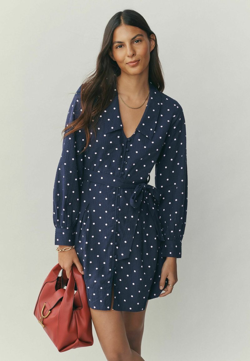 Marineblaues Tupfenkleid mit langen Ärmeln, durchgehender Knopfleiste, V-Ausschnitt, Gürtel in der Taille und Seitenschlitzen, kombiniert mit einer roten Handtasche.