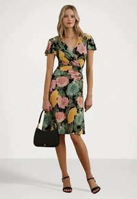 Γυναίκα που φοράει μαύρο φόρεμα μέχρι το γόνατο με μεγάλα ροζ, κίτρινα και πράσινα λουλούδια, κρατώντας μαύρη τσάντα και φορώντας μαύρες γόβες με λουράκι στον αστράγαλο.