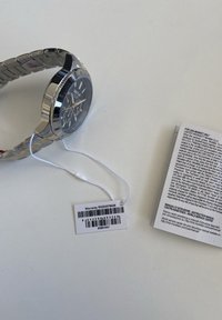 Orologio in acciaio inossidabile argentato con quadrante nero, dotato di cronografo e un'etichetta di garanzia bianca allegata. Manuale di istruzioni visibile accanto ad esso.