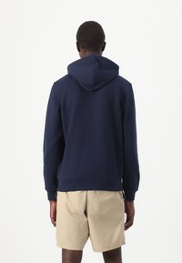 Felpa navy blue in cotone con cappuccio regolabile, polsini a coste e orlo, abbinata a pantaloni corti beige chiaro.