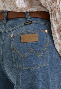 Μπλε τσέπη τζιν Wrangler με καφέ δερμάτινη ετικέτα, κίτρινη ραφή που σχηματίζει ένα "W,", φορεμένη με καφέ ζώνη και τοπ με floral σχέδιο.