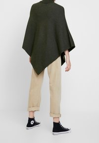 Ciemnozielone dziergane ponczo z wysokim kołnierzem, zestawione z beżowymi spodniami z podwiniętymi nogawkami i czarnymi wysokimi trampkami. Gładka faktura, oversize'owy krój.