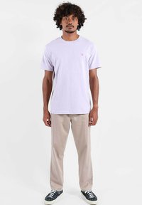 T-shirt en coton violet clair à manches courtes avec un petit logo rose sur la poitrine, associé à un pantalon beige et des baskets noires.