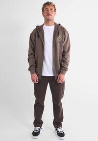 Felpa marrone con zip e grafica bianca, indossata sopra una t-shirt bianca, abbinata a pantaloni marroni e sneakers nere, in piedi davanti a uno sfondo semplice.