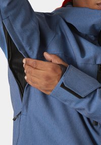 Giacca blu con texture e fodera in rete nera. Presenta una manica parzialmente arrotolata con chiusura in Velcro e un cordino all'orlo.
