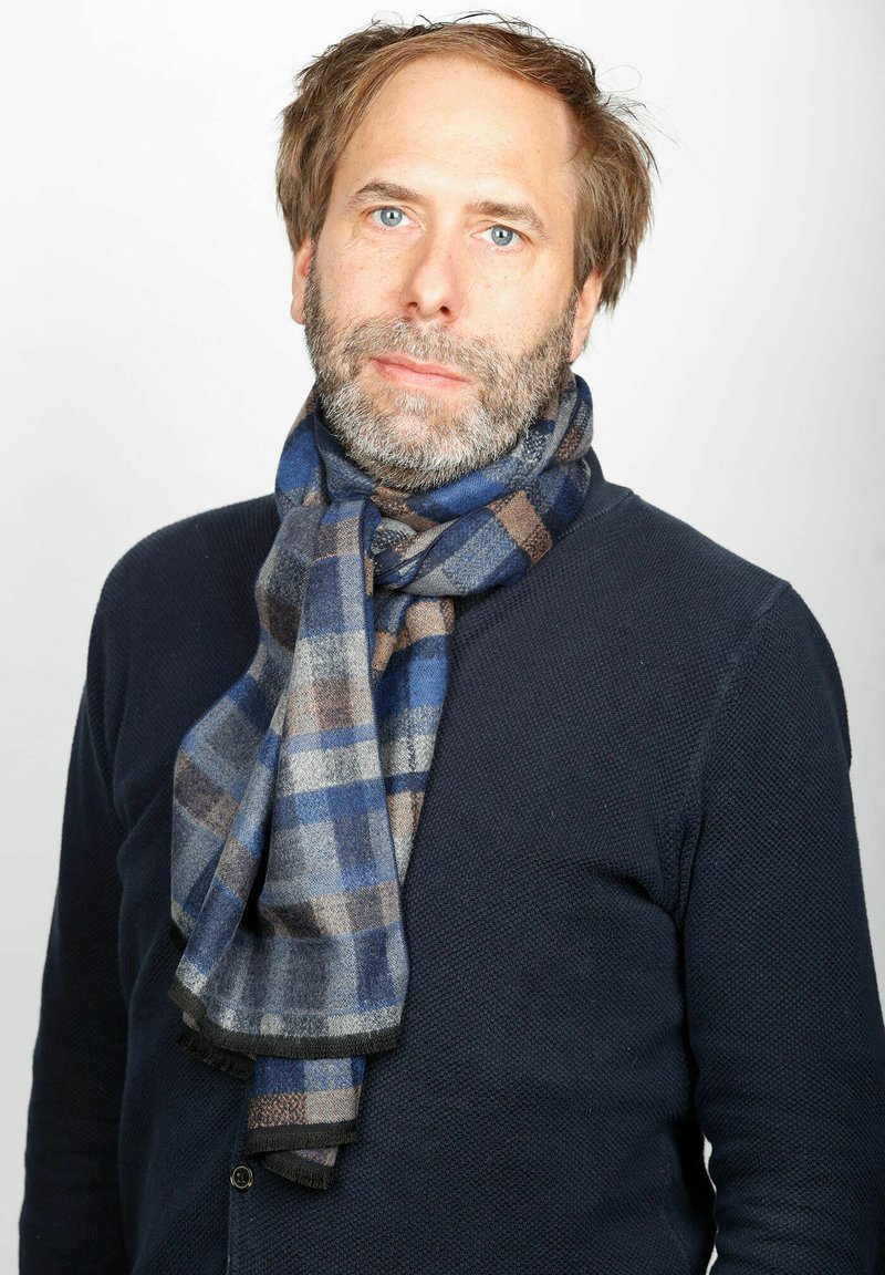 Karierter Schal in Blau, Grau und Braun, um den Hals gewickelt; aus weichem Stoff mit fransigen Kanten, über einem dunklen Pullover getragen.