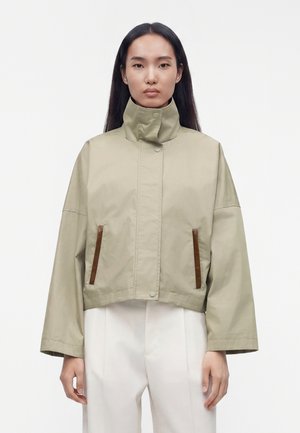 Mujer con chaqueta corta beige con cuello alto y bolsillos con ribete marrón, combinada con pantalones blancos, de pie contra un fondo liso.