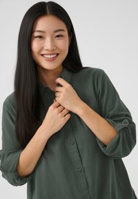 Tmavozelená košeľa na zapínanie s dlhými rukávmi a jemnou textúrou, s golierom a vyhrnutými rukávmi. Modelka sa usmieva a drží golier.