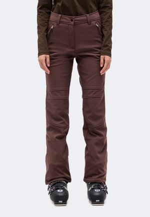 Un mannequin porte un pantalon de ski marron avec des poches zippées et des genoux renforcés. Il est debout, chaussé de bottes de ski, sur un fond uni.