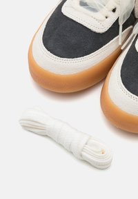 Dark gray suede and cream fabric sneakers with a gum rubber sole, featuring white laces and textured accents. Extra white laces included.