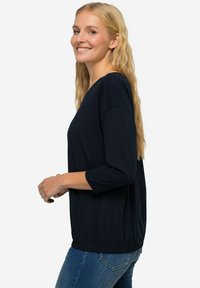 Námořnická modrá blůza s tříčtvrtečními rukávy a nařaseným lemem. Vyrobena z měkké tkaniny, má volný střih a rovnou siluetu.