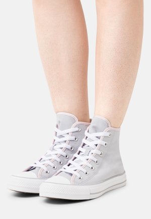 Piernas con zapatillas altas color gris claro con cordones blancos y suelas blancas gruesas, de pie sobre un fondo blanco liso.