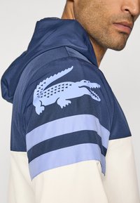 Hooded jack met een blauw en marineblauw kleurenschema, gestructureerde stof, horizontale strepen en een lichtblauw krokodillenlogo op de schouder.
