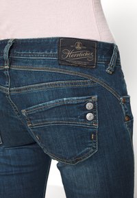 Tummansiniset farkut, joissa on kaksi hopeista nappia takataskussa ja musta kangaslabeli taskun yläpuolella, jossa on kursivoitua bränditekstiä.