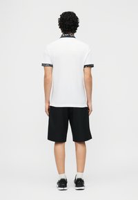 Άνδρας με σγουρά μαλλιά που φοράει λευκή μπλούζα πόλο με μαύρο κείμενο στον γιακά και τα μανίκια, μαύρα σορτς, λευκές κάλτσες και μαύρα αθλητικά παπούτσια, όρθιος με την πλάτη στραμμένη.