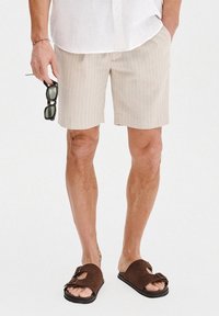 Beżowe spodenki w paski z lekkiego materiału, połączone z brązowymi klapkami. Mężczyzna trzyma ciemne okulary przeciwsłoneczne, ubrany w białą koszulę z guzikami.