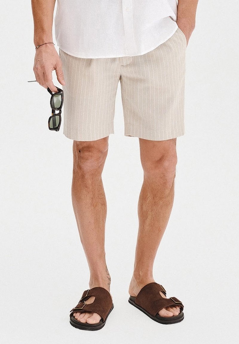 Beżowe spodenki w paski z lekkiego materiału, połączone z brązowymi klapkami. Mężczyzna trzyma ciemne okulary przeciwsłoneczne, ubrany w białą koszulę z guzikami.