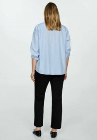 Chemise à boutons rayée bleu clair, avec les manches retroussées, associée à un pantalon noir et des chaussures plates. La chemise a une coupe décontractée et un détail de dos.