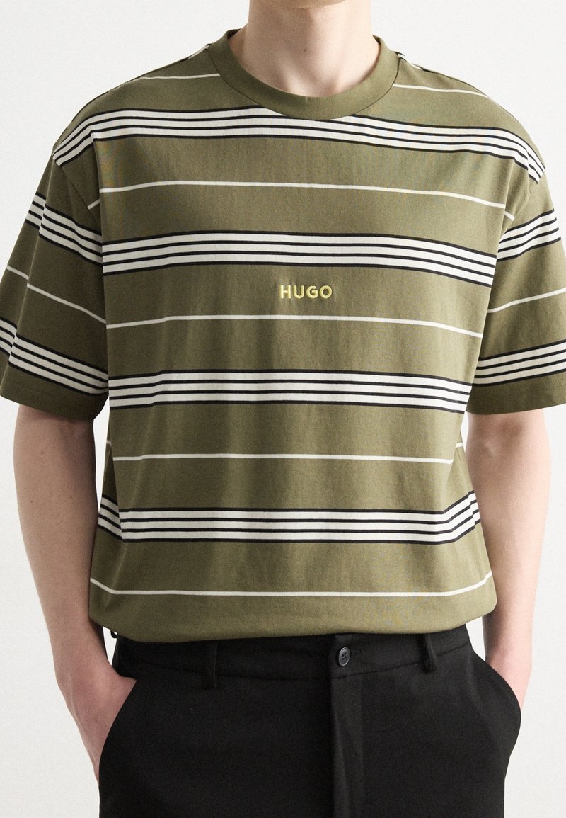 T-shirt rayé vert olive avec des rayures horizontales noires et blanches. Il présente un col rond et le logo « HUGO » en jaune sur la poitrine.