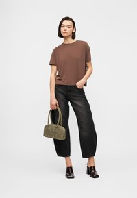 Γυναίκα που φορά καφέ μπλουζάκι και μαύρο τζιν, κρατώντας μια ελιά τσάντα με τρουκς, στέκεται με το ένα χέρι στην τσέπη και φορά μαύρα mules με τακούνι.