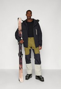Snowboarder portant une veste isolante noire, un pantalon de neige olive et marine, et des bottes noires, tenant un ski avec un design en dégradé.