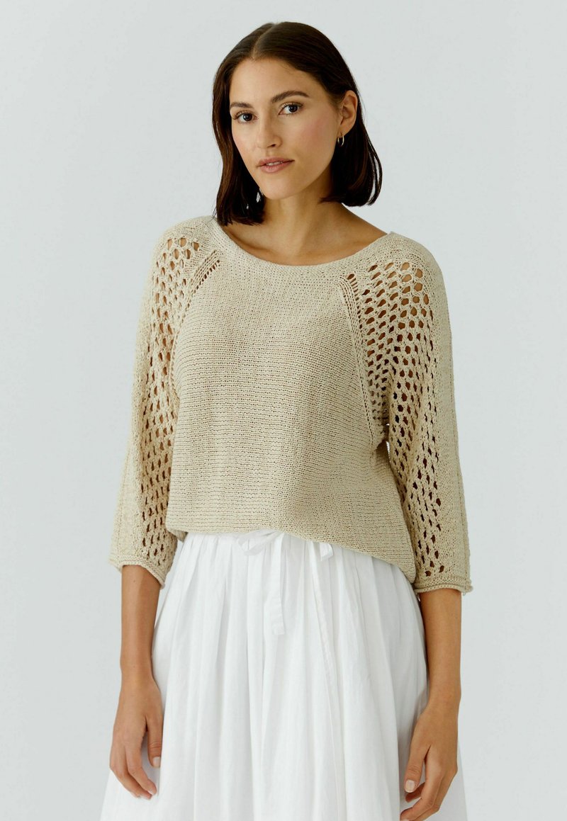 Femme aux cheveux foncés portant un pull beige en maille avec des manches ajourées et une jupe blanche, debout devant un fond clair uni.