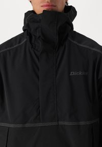 Black windbreaker jacket featuring a high collar, visible white stitching, and a discreet "Dickies" logo on the chest.