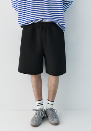 Osoba nosící černé kraťasy po kolena, bílé ponožky s černými pruhy, šedé tenisky a modro-bíle pruhované tričko.