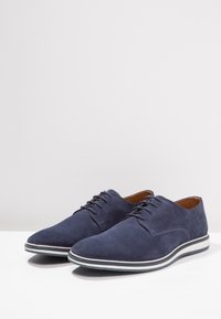 Chaussures habillées en daim bleu marine avec un bout pointu, un design à lacets, une semelle en caoutchouc blanche et une bordure grise contrastante. Texture lisse sur toute la surface.