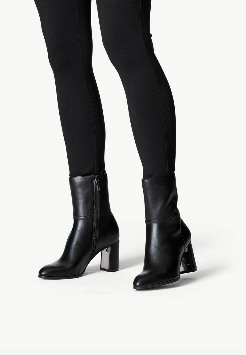 Bottes en cuir noires mi-mollet avec talons blocs et fermetures éclair latérales, portées avec un pantalon noir ajusté, présentées de la moitié du mollet vers le bas sur fond blanc.