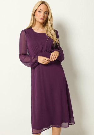 Mujer rubia de pie con las manos entrelazadas, vestida con un vestido morado de manga larga hasta la rodilla con mangas transparentes sobre un fondo claro y sencillo.