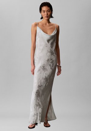 Donna che indossa un vestito a fiori senza maniche color argento-grigio con uno spacco frontale, sandali neri con tacco e un bracciale d'argento al polso destro.