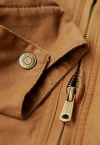 Lichtbruine jas van een gestructureerde stof, met een zichtbare gouden rits en een branded metalen drukknoop als detail.
