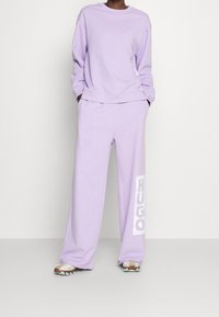 Persona que viste un sudadera lavanda y pantalones de pierna ancha con "HUGO" impreso en una pierna, combinados con zapatillas beige y blancas.