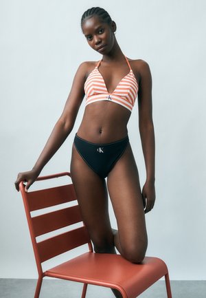 Γυναίκα με πλεξούδες, που φοράει μπικίνι με ριγέ τοπ και μαύρο κάτω μέρος, στηριζόμενη σε κόκκινη καρέκλα μπροστά σε μονόχρωμο φόντο.