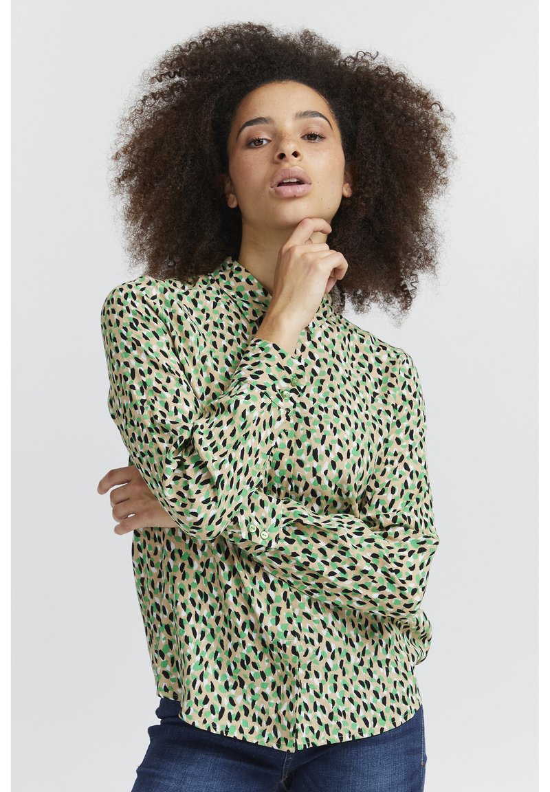 Blusa a maniche lunghe con colletto di colore verde chiaro, decorata con un motivo di foglie nere e verdi sparse. Tessuto morbido e leggero, con polsini non abbottonati.