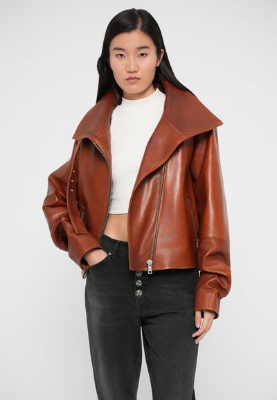 Chaqueta de cuero marrón con un collar ancho, diseño corto y cremalleras plateadas. Se lleva sobre una blusa blanca, combinada con jeans negros de cintura alta.