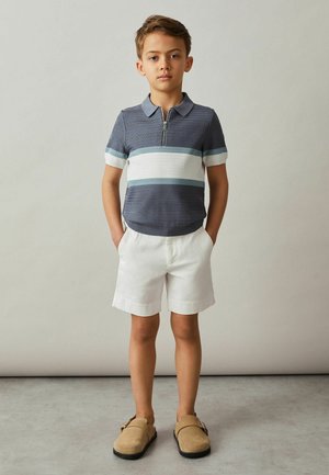 Jongen staat met handen in de zakken, draagt een blauw gestreept poloshirt, witte shorts en beige instapschoenen tegen een effen achtergrond.